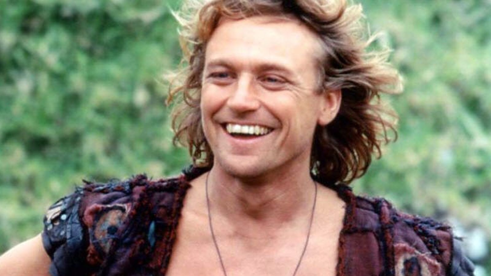 a photo of a middle aged white man with long light blonde hair wearing a vest and a jade necklace
