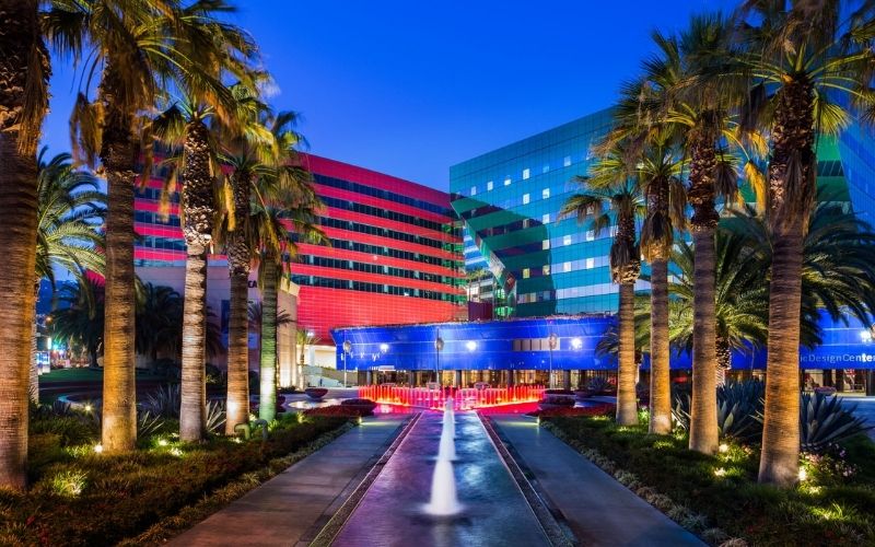 Vibrant hotel exterior at dusk with palm trees lining a fountain path. Blue and red lights illuminate modern glass buildings, creating an inviting ambience.