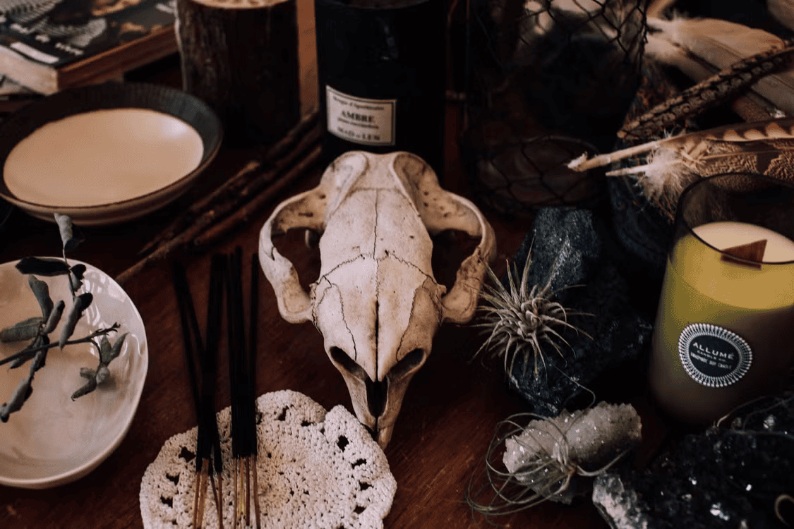 Domestic Rituals A skull rests on a wooden table surrounded by candles, feathers, crystals, incense sticks, dishes, a crocheted doily, and small plants.