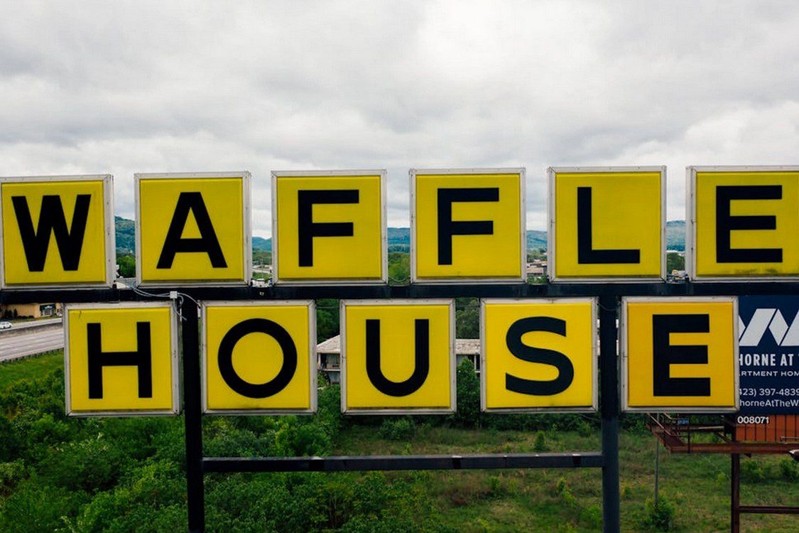 wafflehouse-2 Yellow Waffle House sign with black letters stands outdoors against a cloudy sky and greenery in the background.