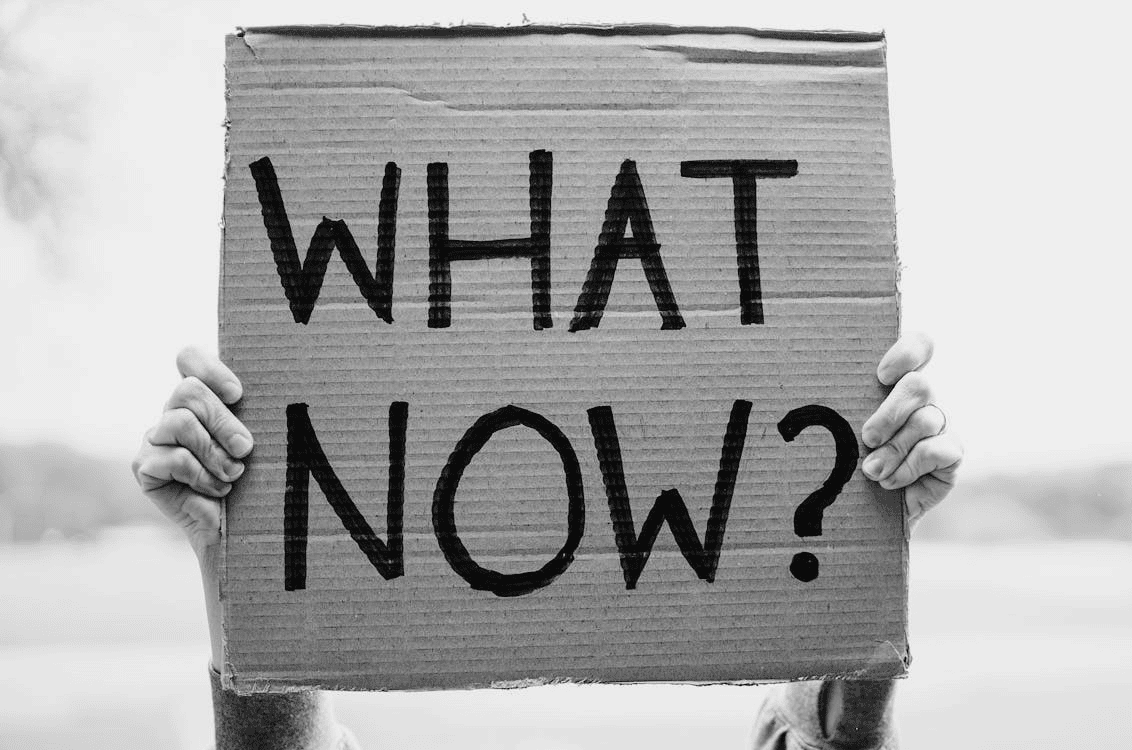 Black-and-white photo of hands holding a cardboard sign reading “WHAT NOW?” in bold letters. Black-and-white photo of hands holding a cardboard sign reading “WHAT NOW?” in bold letters.